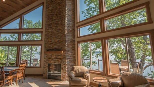 Amplia habitación con chimenea de piedra, ventanas grandes con vistas a árboles y un lago, y un techo alto con vigas de madera.