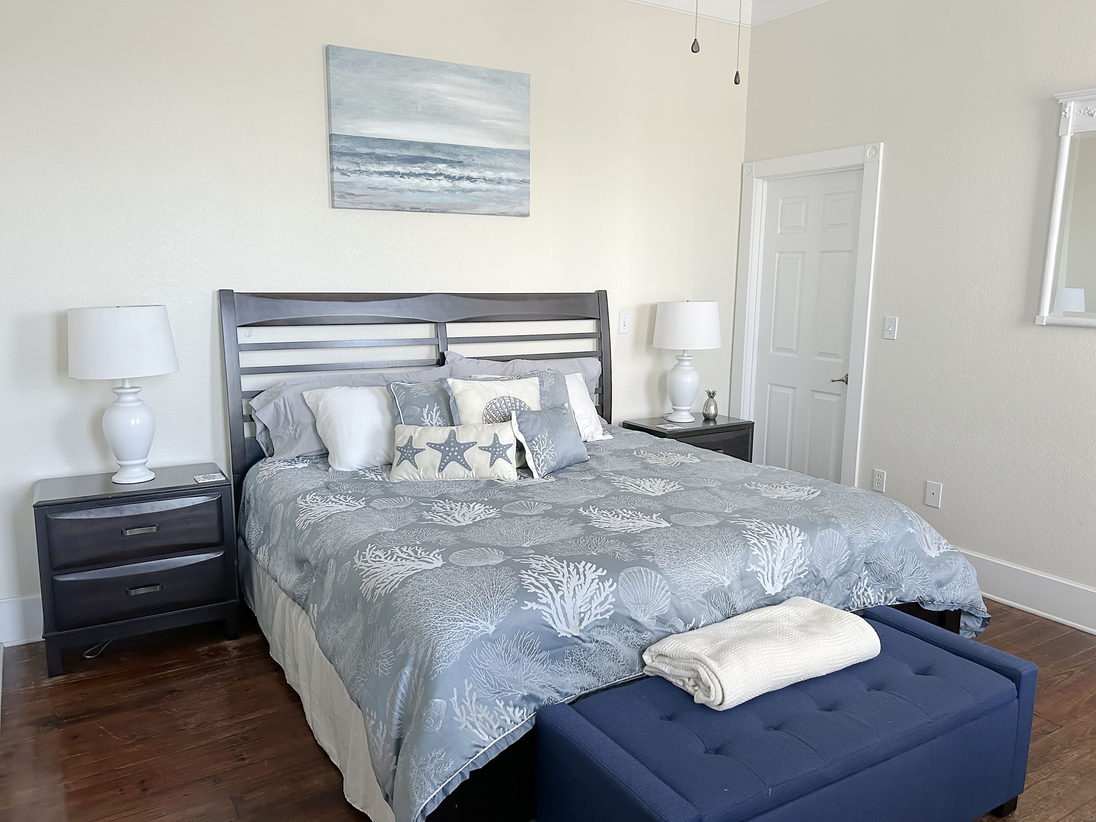 Un dormitorio ordenado con cama, dos mesitas de noche, un banco y un cuadro en la pared.
