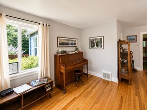 A room with a wooden piano, a window with curtains, a bookshelf, and framed artwork on the wall. - CUTE and COMFORTABLE, in a quaint location, Central AC, DOG friendly (Hood River)