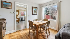 Uma área de refeições com uma mesa de madeira e cadeiras de vime, uma janela com cortinas, e vista para árvores.