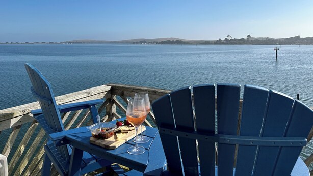 Una mesa de comedor con una copa de vino, un cuenco de fruta y una tabla de quesos en una terraza de madera con vistas al agua.