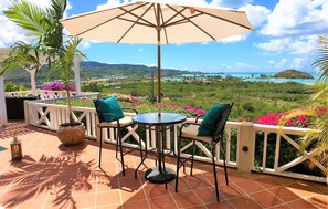 Een terras met een tafel, stoelen, een parasol en uitzicht op een kustlandschap.