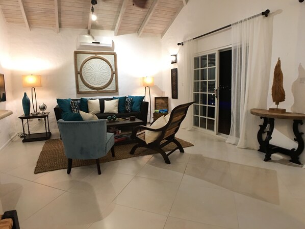 Un salon comprenant un canapé, des fauteuils, une table basse et une cheminée. Il y a une grande fenêtre avec des rideaux, une porte et un climatiseur fixé au mur.