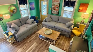 A living room with two grey sofas, a wooden coffee table, and a yellow armchair. There are two windows with curtains, a ceiling fan, and a wall with a poster. - Four Blocks To Beach & Boardwalk  + Pet Friendly (Ocean City)