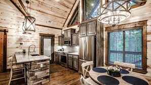 A rustic cabin interior with a stone island, wooden dining table, and a large window with blinds. - Elegant Couples Cabin, Magnolia Design, 5-Star Amenities (Broken Bow)