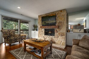 A living room with a stone fireplace, a television mounted above it, and a large window offering a view of trees.