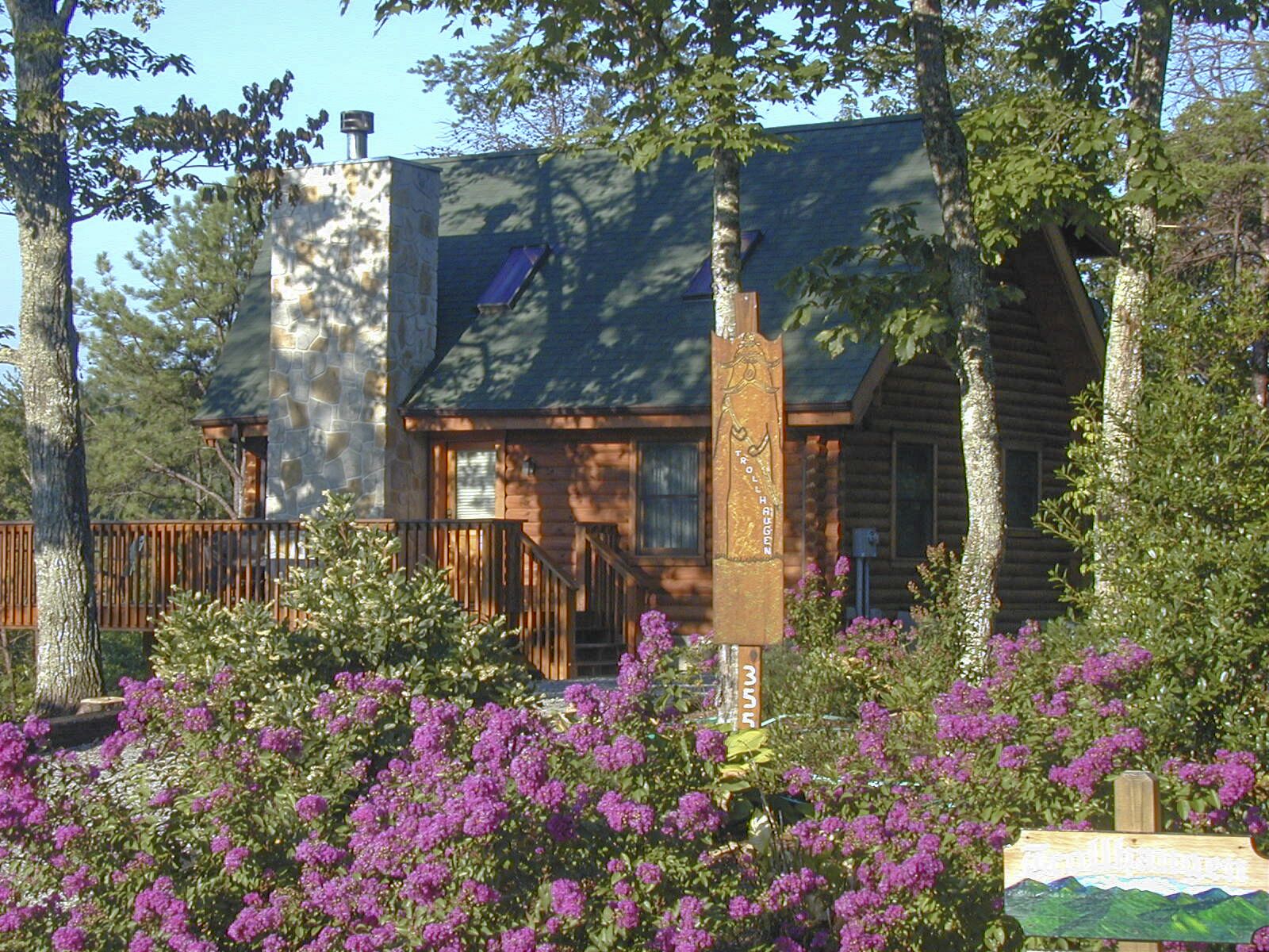 A log cabin with a stone chimney, surrounded by lush greenery and purple flowers.