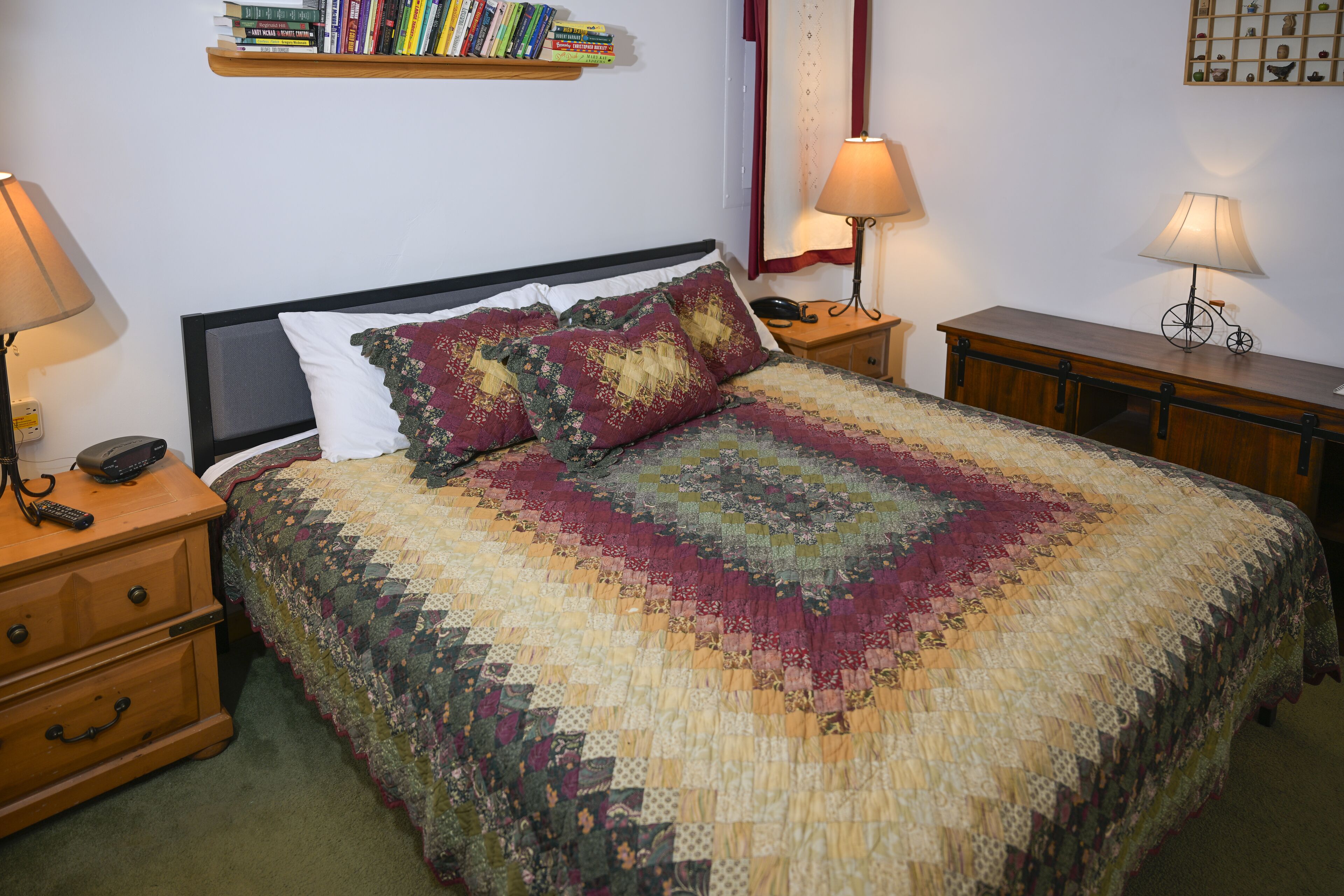 A neatly made bed with a quilt, two bedside tables with lamps, a nightstand, and a bookshelf with books.