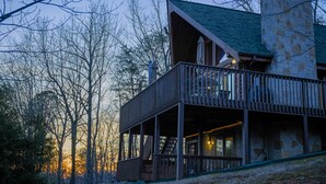 A multi-level wooden cabin with a stone chimney, a balcony with outdoor seating, and a view of the surrounding trees. - Trollhaugen—Enjoy Spring and Summer in the Great Smoky Mountains (Sevierville)