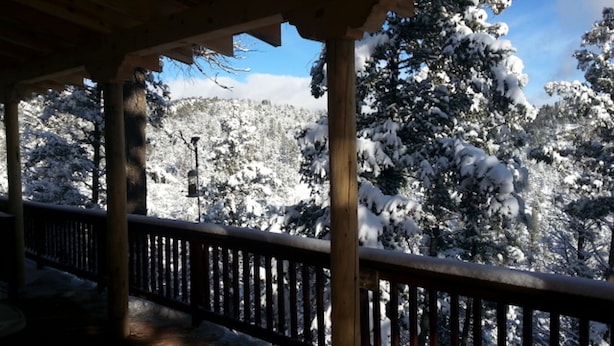 A wooden deck with a railing, overlooking a snowy landscape with trees and a clear sky.