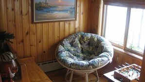 Quarto com paredes de madeira, um quadro de uma paisagem costeira, uma cadeira redonda com almofada florida, uma mesa de futebol de mesa e uma janela com vista.
