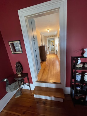 A hallway with wooden flooring, a staircase leading to another room, and a shelf with decorative items.