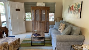 A living room with a grey sofa, a wooden cabinet with a 'welcome' sign, a patterned rug, and a framed artwork on the wall.