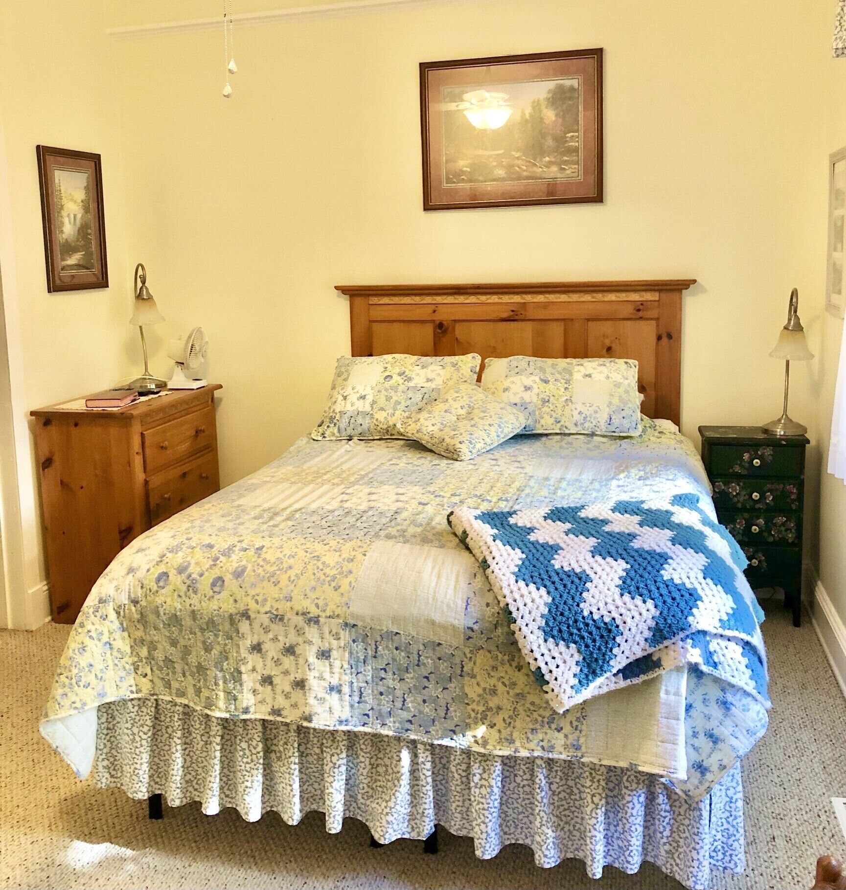Un dormitorio con cabecera de madera, ropa de cama floral, colcha, mesita de noche con lámpara y cómoda.