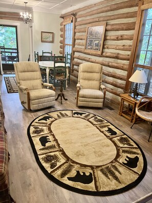 A log cabin interior with two beige armchairs, a wooden dining table, chairs, a chandelier, and a rug with animal prints.