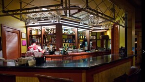 Zona de bar con estantes llenos de botellas de licor, un bartender y copas colgantes.
