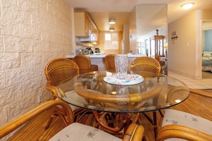 A dining area with a glass table surrounded by wicker chairs, a kitchen in the background, and a brick wall.