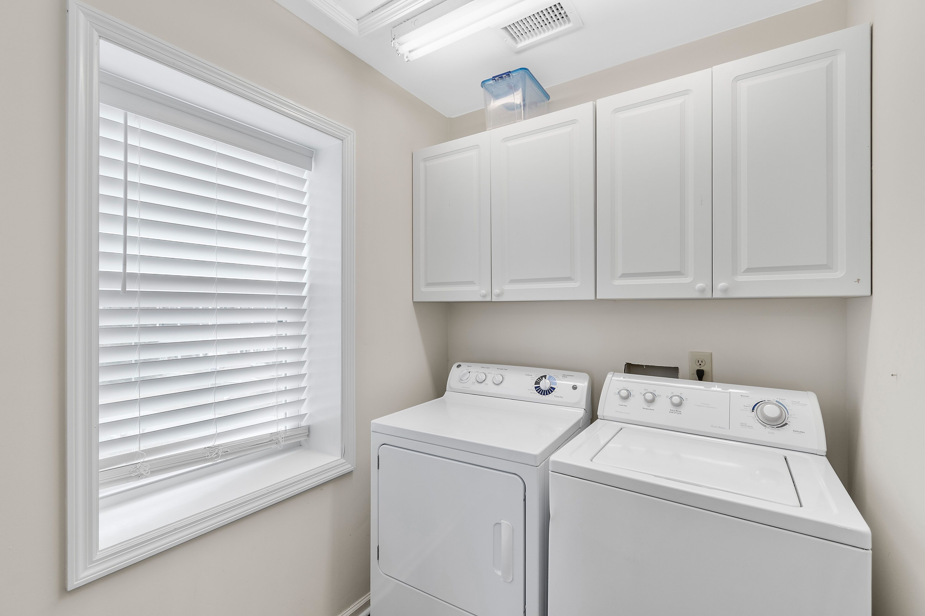 A laundry room with two white front-loading washers and dryers, a window with blinds, and a ceiling vent.