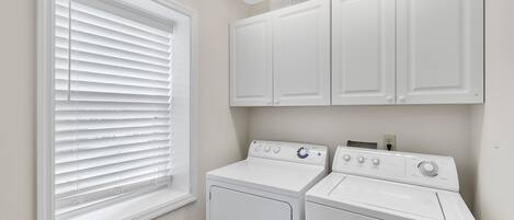 A laundry room with two white front-loading washers and dryers, a window with blinds, and a ceiling vent.