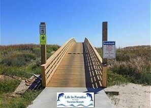 Une promenade en bois mène à une plage, et un panneau indique une limite de vitesse de 5 miles par heure.