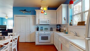 A kitchen with white cabinets, a microwave, an oven, and a sink. There is a ceiling fan, a fish decoration, and a sign on the window. - Golf Cart Included, Shared Pool, Pet Friendly, Boardwalk to Beach (Port Aransas)