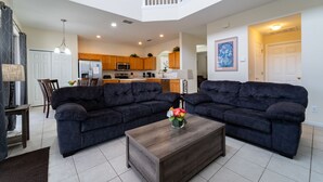 A spacious living room with two dark sofas, a wooden coffee table, and a dining area with a table and chairs. A kitchen is visible in the background with wooden cabinets and a microwave.