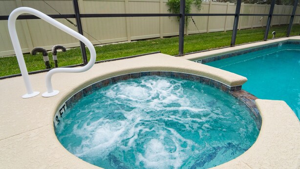A hot tub with a 3 FT depth marker, surrounded by a pool and a grassy area with a fence in the background.