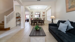 A spacious living room with a dark wood coffee table, a black sofa with decorative pillows, a staircase with white railings, and a dining area with a chandelier.