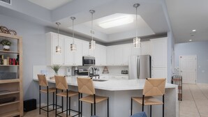 Una cocina moderna con armarios blancos, una isla central con taburetes de bar, electrodomésticos de acero inoxidable e iluminación colgante.