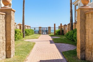 A paved pathway leading to a gated entrance with a clear view of the sea in the background.