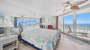 A hotel room with a bed, a desk, a chair, a balcony with a view of the ocean, and a ceiling fan.