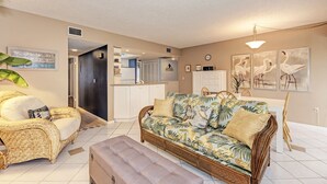 Ein Wohnzimmer mit einer Couch, einem Sessel und einem Esstisch. Im Hintergrund ist eine Küche zu sehen.