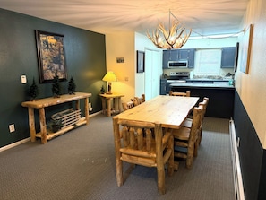 A dining area with a wooden table and chairs, a chandelier, a small lamp, and a kitchen in the background.