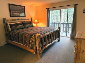 A bed with a patterned bedspread, a wooden headboard, a nightstand with a lamp, and a view of trees through a large window.