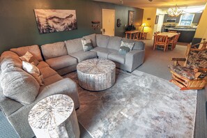 A living room with a sectional sofa, ottomans, a dining table with chairs, and a kitchen in the background.