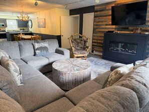 A living room with a sectional sofa, a round coffee table, a fireplace, and a television mounted on a wooden wall.