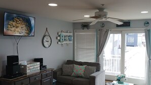 Una sala de estar con televisor, reloj, ventilador de techo y una ventana con cortinas.