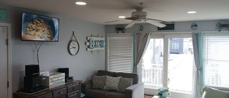 A living room with a television, clock, ceiling fan, and a window with curtains.