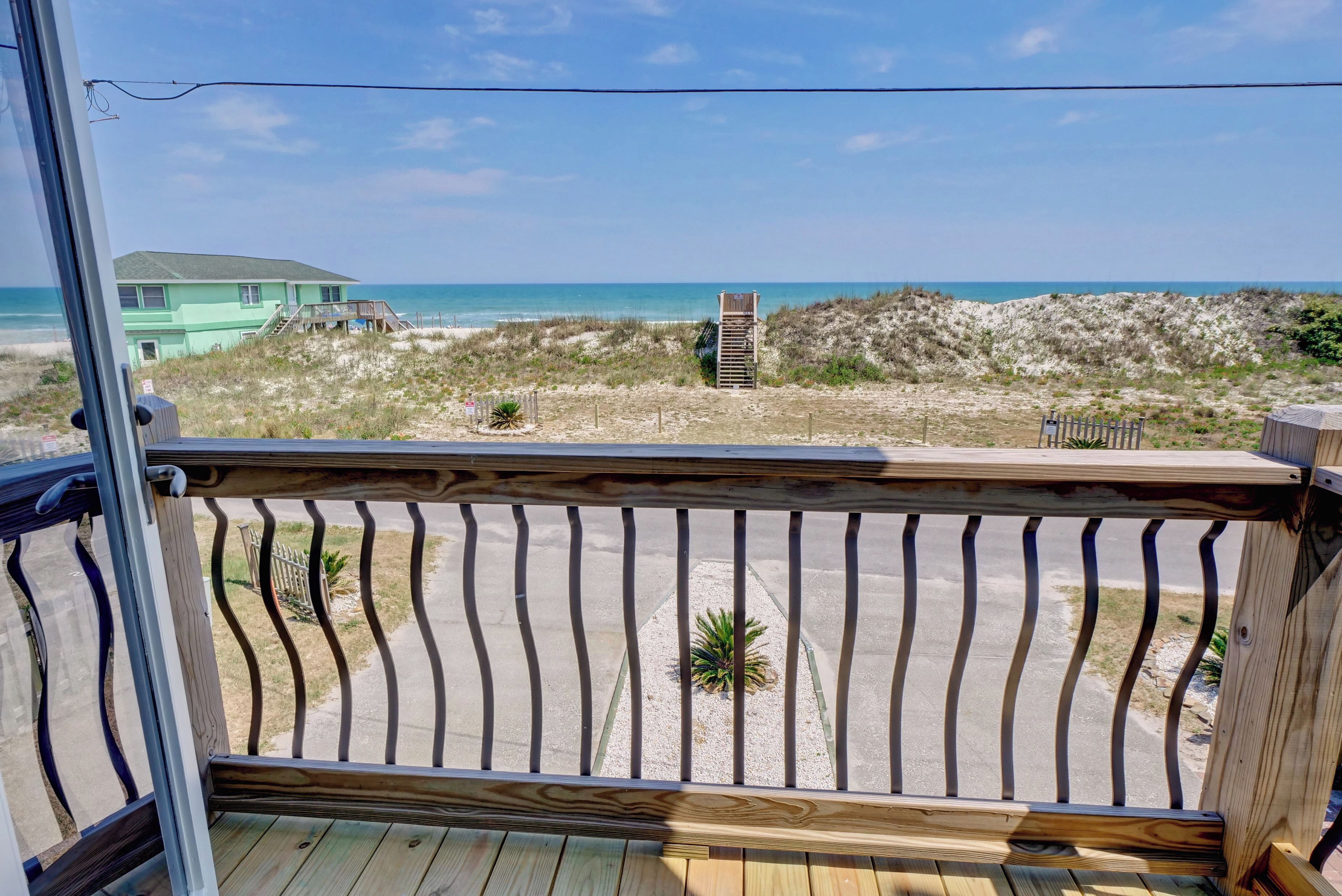 Une terrasse en bois avec une rambarde en métal, donnant sur une plage avec une dune et une maison en arrière-plan.