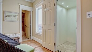 Una habitación con una cama, un banco tapizado de color verde, una obra de arte enmarcada, una puerta que da al baño y un suelo de baldosas.