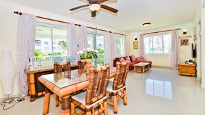 A spacious living room with a dining area, large windows, and a ceiling fan. - Playa Turquesa Luxury Beach Front Condo with Ocean View and Much More (Bávaro)