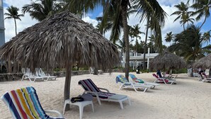 A beach with white lounge chairs, some with colorful towels, and thatched umbrellas. - Playa Turquesa Luxury Beach Front Condo with Ocean View and Much More (Bávaro)