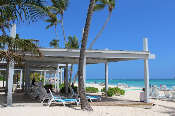 A beachside lounge with white lounge chairs and a covered seating area, overlooking a sandy beach and the ocean.