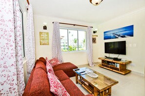 A living room with a sofa, a coffee table, a TV, and a window with curtains. - Playa Turquesa Luxury Beach Front Condo with Ocean View and Much More (Bávaro)