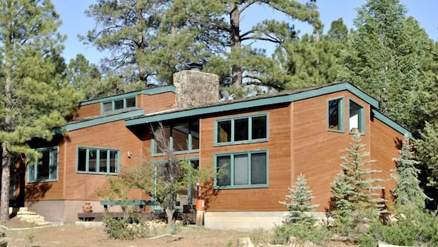 Una casa moderna de dos pisos con fachada de madera y detalles verdes, rodeada de árboles y un paisaje seco.