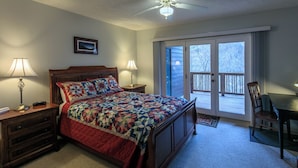 A bedroom with a large bed, two bedside lamps, a nightstand, a wooden dresser, a chair, a dining table, and a balcony with a view of trees.
