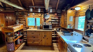 A rustic cabin kitchen with wooden cabinets, a central island, a stove, and a window overlooking greenery. - Blinn's Landing: Historic Riverfront Log Cabin, Dock & Mountain Views. Dogs OK! (Bend)