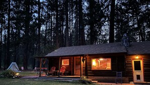 A log cabin with a porch, a dining area, and a visible staircase leading to the upper level. - Blinn's Landing: Historic Riverfront Log Cabin, Dock & Mountain Views. Dogs OK! (Bend)
