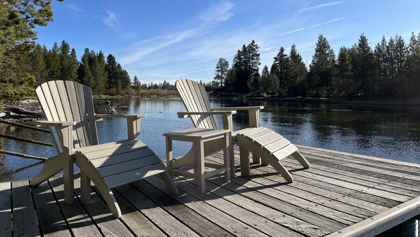 A wooden deck with two Adirondack chairs facing a calm lake surrounded by trees. - Blinn's Landing: Historic Riverfront Log Cabin, Dock & Mountain Views. Dogs OK! (Bend)