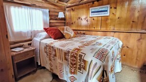 A wooden cabin room with a bed, a nightstand, a lamp, and a framed picture on the wall. - Blinn's Landing: Historic Riverfront Log Cabin, Dock & Mountain Views. Dogs OK! (Bend)